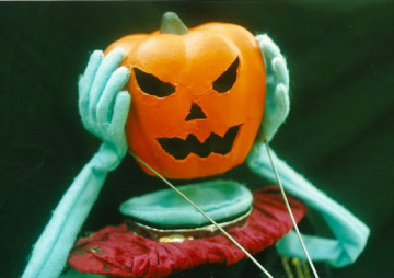 a puppet with a carved orange jack o lantern head holding its hands to the sides of its face.