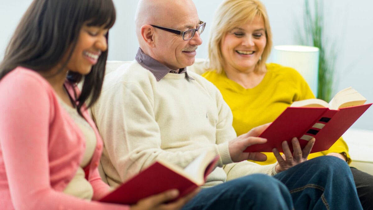 three adults sit together on a couch, smiling and reading books with red covers. the atmosphere appears relaxed and friendly, with soft indoor lighting in the background.