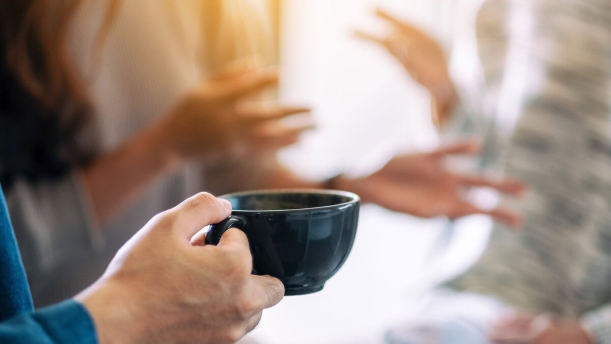 Close-up of a person holding a black mug, with two other people in the background gesturing as they have a conversation. The scene suggests a casual, social interaction, possibly in a home or office setting.