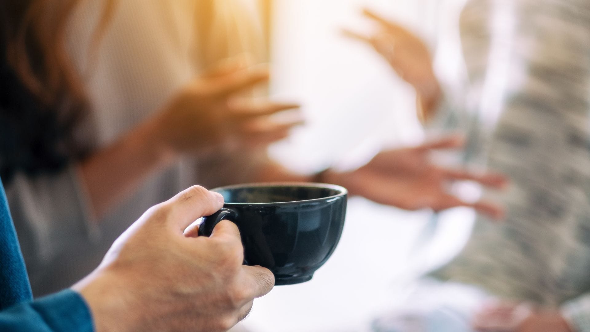 Close-up of a person holding a black mug, with two other people in the background gesturing as they have a conversation. The scene suggests a casual, social interaction, possibly in a home or office setting.