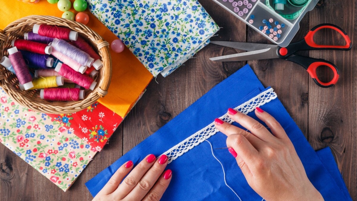 A person sews white lace onto blue fabric; nearby are colorful fabric pieces, thread spools in a basket, buttons, a sewing kit, and red-handled scissors on a wooden table.