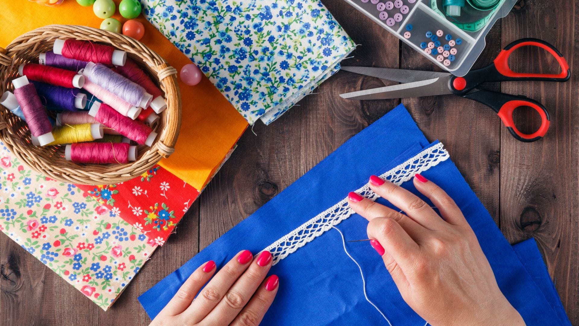 A person sews white lace onto blue fabric. Nearby are floral fabrics, a basket of colorful thread spools, scissors, and a box of sewing supplies, all on a wooden table.