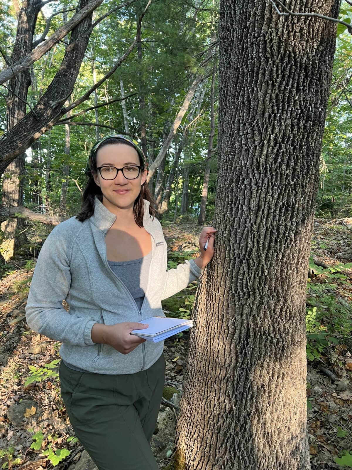 a person with glasses stands in a sunlit forest, holding a notebook and pen, leaning against a tree with textured bark. green leaves and dappled sunlight surround them.