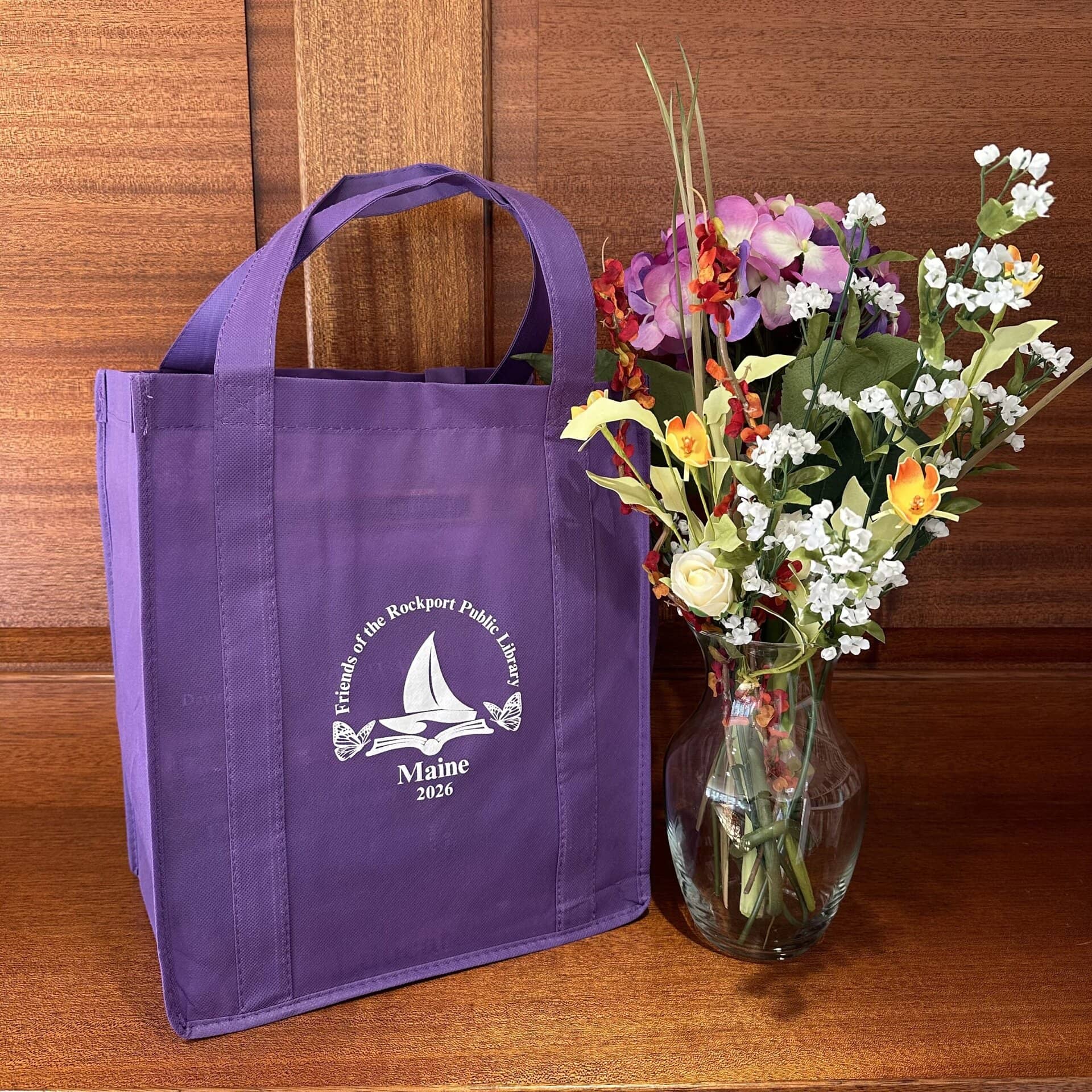 a purple reusable tote bag with a white sailboat graphic and friends of the rockport public library maine 2026 sits next to a vase of colorful artificial flowers on a wooden surface.