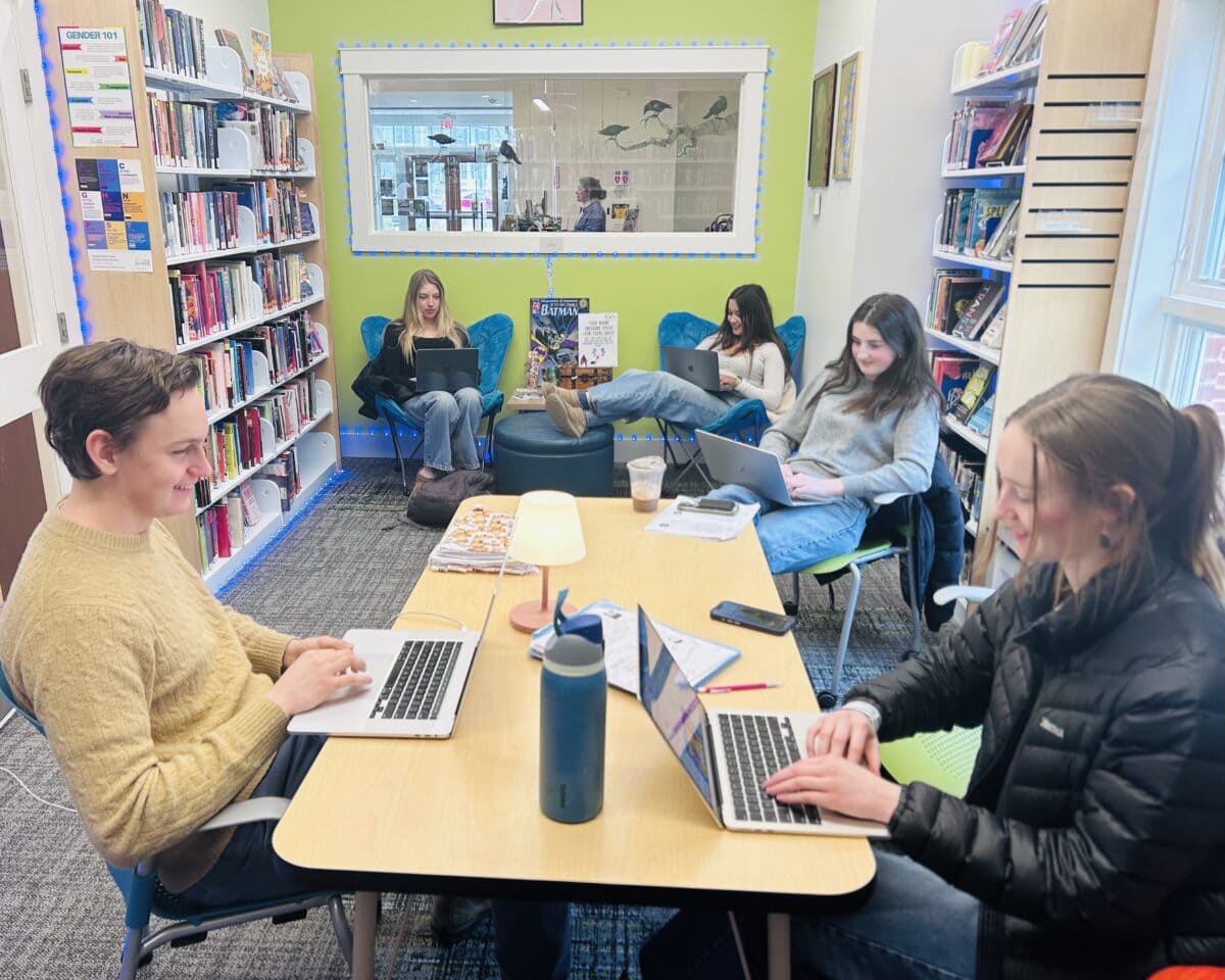 five people work on laptops in a bright library room, surrounded by bookshelves. four are at a table, while one sits on a blue chair in the background. a lamp and water bottle are on the table.