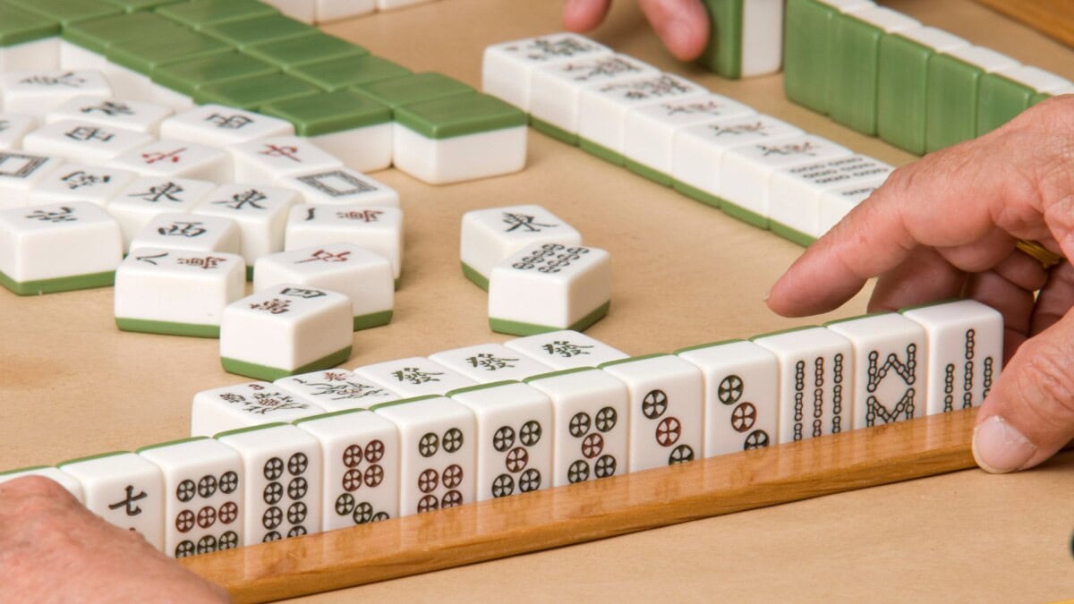 Close-up of people playing mahjong, with hands arranging white and green mahjong tiles on a wooden rack and game table. Some tiles have Chinese characters and symbols.