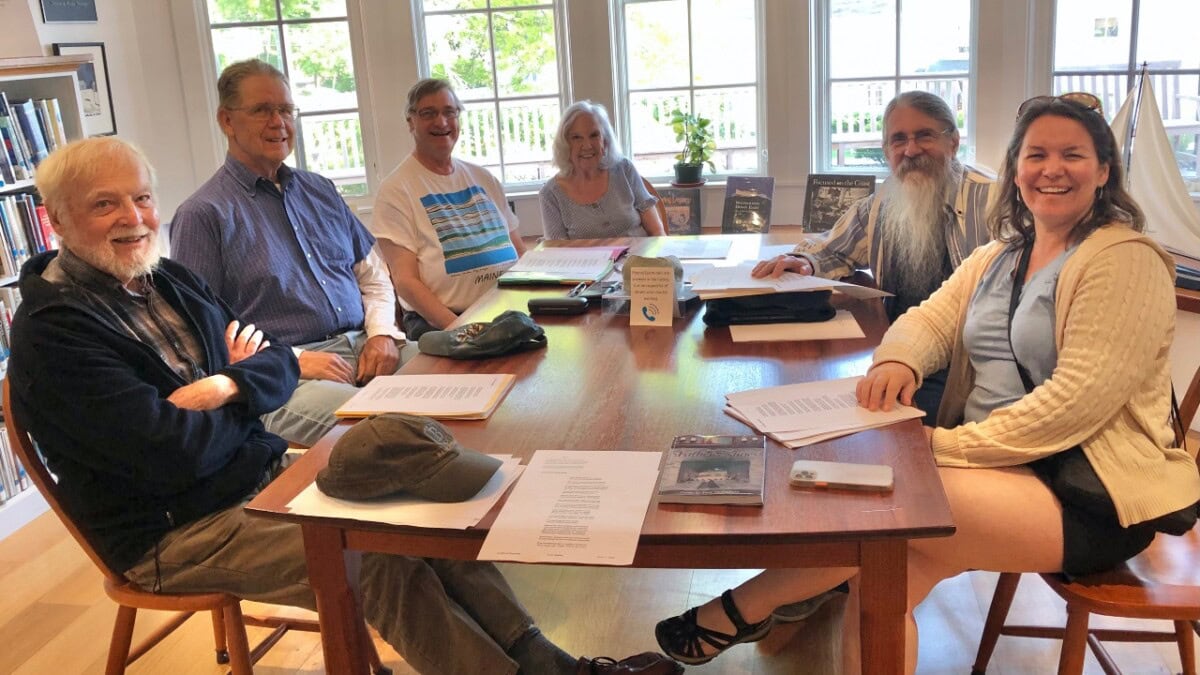Six smiling adults, four men and two women, sit around a wooden table in a bright room with large windows. Papers, books, and a hat are on the table; a model sailboat is visible on the right.