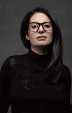 A woman with long dark hair and glasses wears a black turtleneck sweater and looks confidently at the camera against a dark gray background.