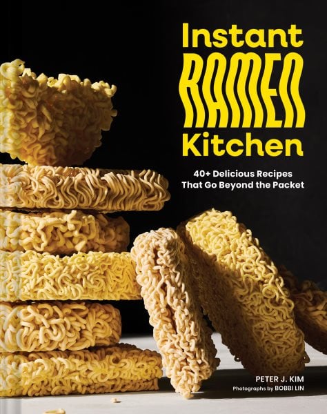 stacks of uncooked instant ramen noodle blocks are arranged in front of a black background. the book title instant ramen kitchen and subtitle appear in bold yellow and white text on the right side.