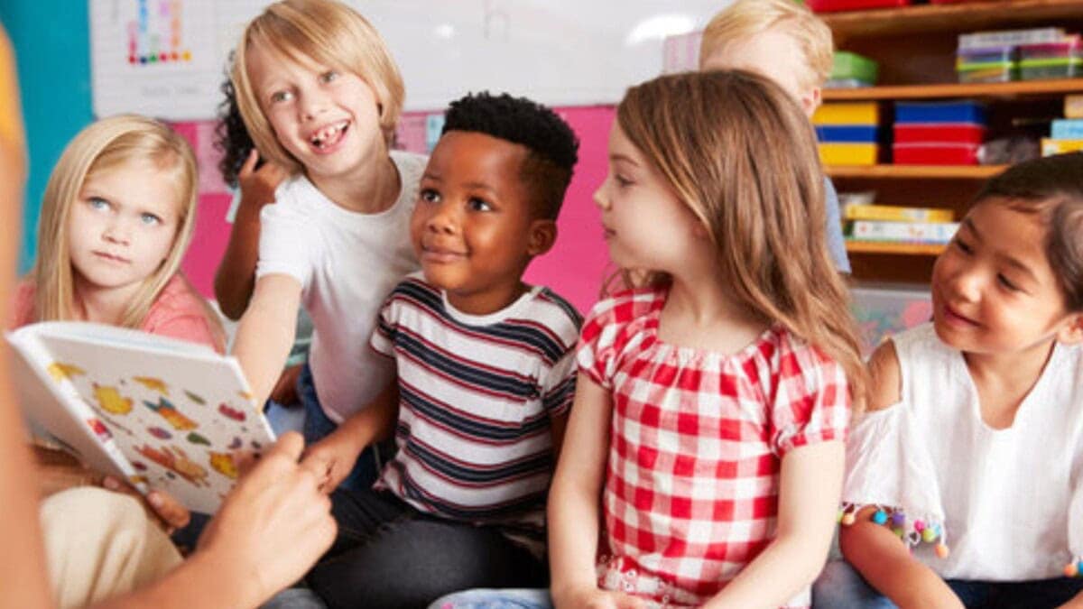 a group of young children sit closely together on the floor, smiling and listening as an adult reads aloud from a picture book in a colorful classroom.