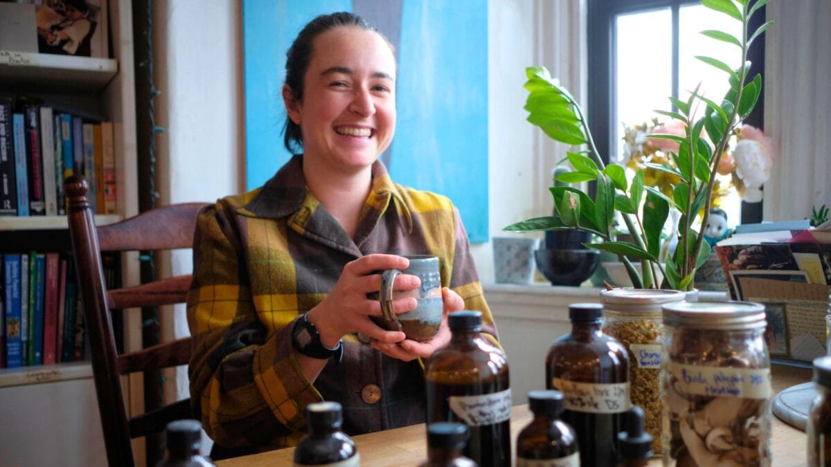 a smiling person in a plaid jacket holds a mug at a table with glass jars and bottles. shelves with books and a green potted plant are visible in the background near a window.