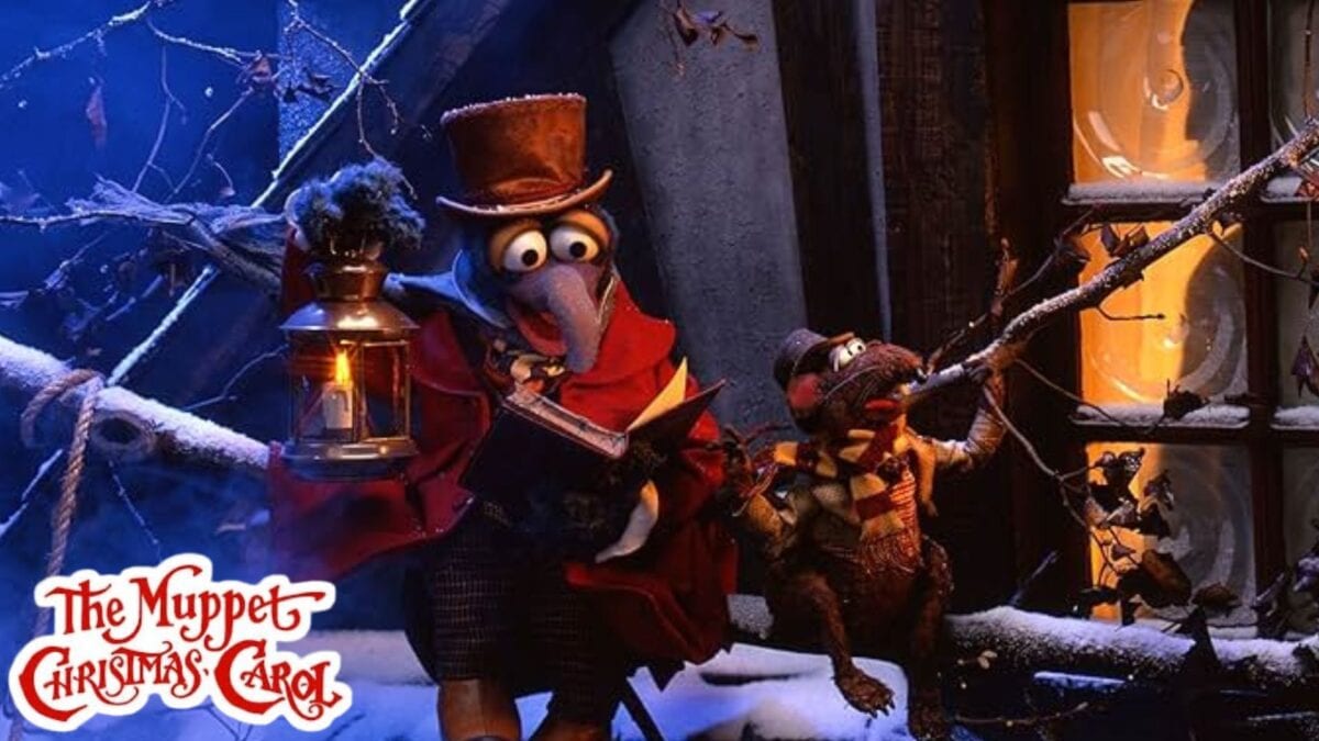 gonzo and rizzo the rat, dressed in victorian winter clothes, stand outside a snowy window at night. gonzo holds a lantern and a book. the “the muppet christmas carol” logo is in the bottom left corner.