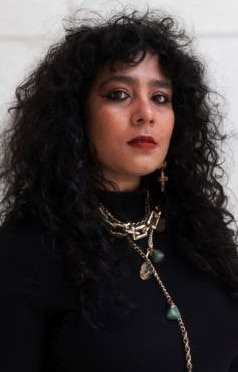 A woman with long, curly black hair wearing a black top, layered gold necklaces, and bold makeup with dark eyeliner and red lipstick, stands against a light-colored background.